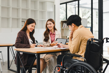 Young businesswoman meeting client in wheelchair, smiling colleagues discuss documents and tablet in modern office, inclusive teamwork and support
