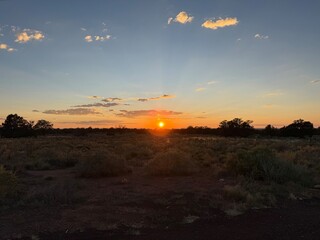 Dramatic Desert Sunset: Intense red and orange sun rays pierce a gradient sky, transitioning to deep blue, silhouetting a distant city skyline and arid desert bushes.