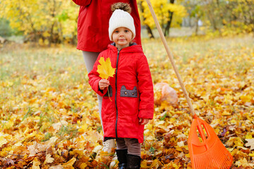 A young child in a red coat holds a yellow leaf while standing next to a parent raking leaves in a vibrant autumn park