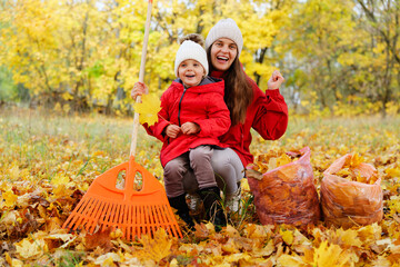 A mother and her young daughter smile happily while raking leaves on a crisp fall day, surrounded by golden trees in the park