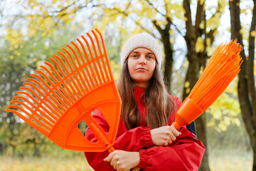 Woman dressed in warm clothing stands in a forest holding an orange rake and broom, ready for autumn yard work amid colorful leaves