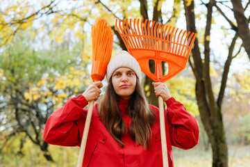 A young woman dressed in a red jacket and white hat stands in a park, holding orange rakes and looking determined among autumn foliage