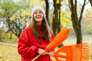 In a park filled with colorful autumn leaves, a happy woman holds orange rakes while wearing a warm red jacket and a cozy hat