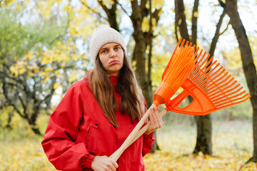 A young person stands in a colorful autumn forest, holding an orange rake while looking serious