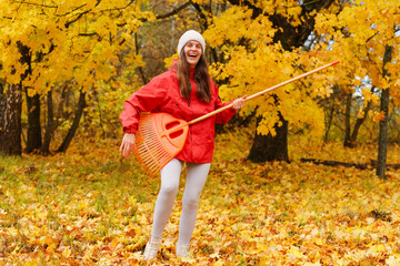 A woman is smiling and raking colorful leaves in a forest during autumn. The scene is filled with bright yellow trees