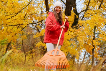 A young woman rakes fallen leaves in a vibrant autumn forest, wearing a cozy red jacket and a beanie, enjoying the beautiful weather