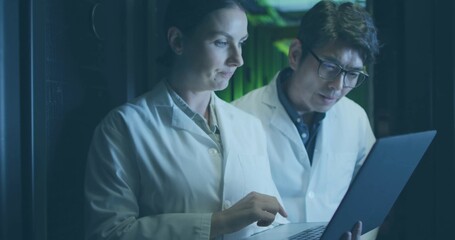 Technicians in lab coats tapping laptop while colleague checking server racks in aisle, green LEDs