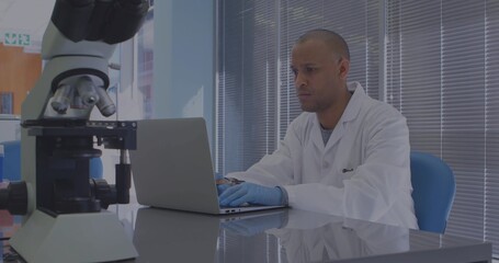 Typing scientist wearing lab coat and gloves entering data on laptop in lab, with microscope