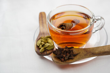 Aromatic black tea or red tea served hot in a glass cup. Spices like cardamom, cinnamon and cloves, are scattered around. Healthy traditional drink, side view and selective focus.