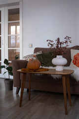 This cozy living room with a pumpkin on wooden table, plants in pot, sec autumn branches in ceramic vase and modern furnishings creates an inviting autumn atmosphere.
