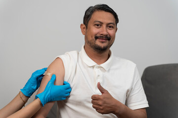 Smiling man showing thumb up after vaccination with adhesive bandage on arm receiving healthcare service
