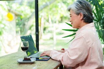 Senior Asian woman focused laptop cafe casual clothing working drinking wine dessert plate relaxing indoor natural light atmosphere leisure lifestyle refreshing vacation freedom happiness cheerful