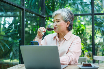 Senior Asian woman sipping red wine smiling working laptop cafe casual outfit greenery indoor background relaxing leisure communication freelance lifestyle freedom happiness retirement business