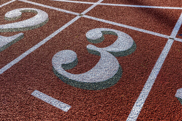 Close up photo of outdoor running track lane white number three, 3, with green shadow, on a new red track with white lane lines.