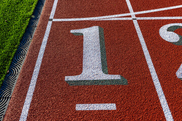 Close up photo of outdoor running track lane white number one, 1, with green shadow, on a new red track with white lane lines.