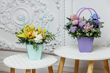 Two contrasting small flower basket arrangements one turquoise with orchids and one purple with cotton displayed on white decorative tables in a bright interior