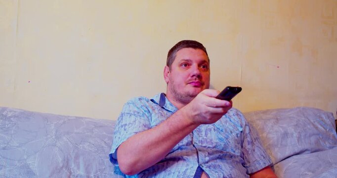 Tired middle-aged man sitting on couch holding TV remote, looking exhausted after long day at home.