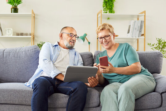 Elderly couple on sofa at home share a tablet and smartphone, browsing internet for social media and explore apps together. Seniors learns online banking in retirement. Leisure and technology. 