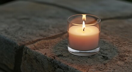 Lit candle in glass holder on textured stone surface with soft light votive candle