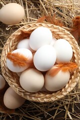 Raw chicken eggs in wicker basket, feathers and straw on table, top view