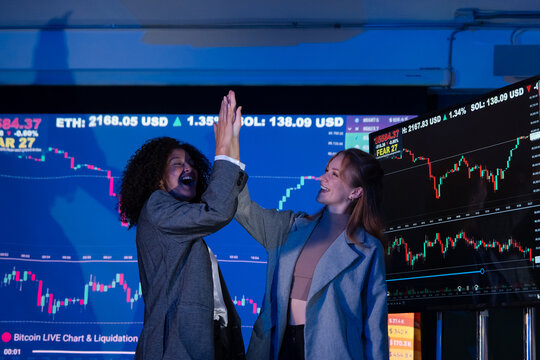 Two excited female traders celebrating a successful investment, raising arms in victory in front of stock charts.