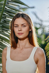 Young woman with long brown hair and natural makeup stands outdoors near palm leaves, wearing a...