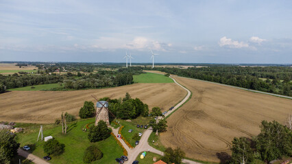 Aerial view of a rural landscape with an old stone windmill, winding road, farmlands, green fields, and wind turbines under a clear blue sky