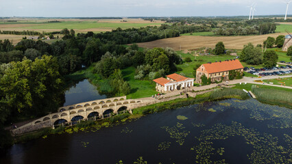 Obraz premium Aerial view of a historic stone bridge over a calm river with red-roofed buildings, green fields, and wind turbines in the background