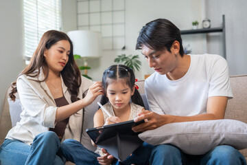 Young parents and daughter sitting on couch, learning and bonding while drawing on a digital tablet in the living room