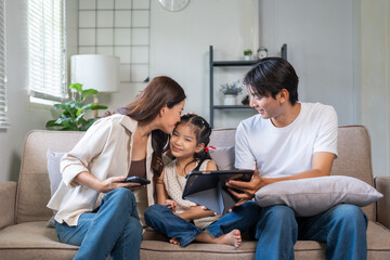 Asian parents and child enjoying time together on living room sofa, mother kissing daughter, father using tablet