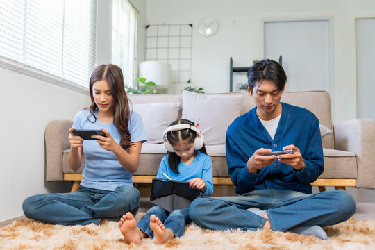 Asian family sitting on the floor, mother and father playing games on smartphones, daughter drawing on digital tablet with headphones