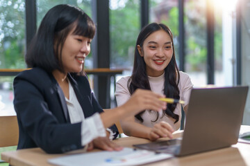 Two smiling women are collaborating at a desk, reviewing data on a laptop and discussing business ideas in a modern office