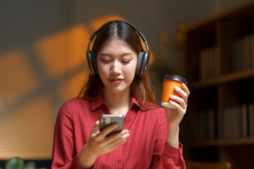 Young Asian woman wearing headphones, holding a coffee cup, and looking at a smartphone, listening to music and relaxing