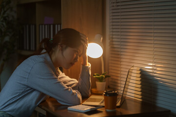 Asian woman experiencing burnout and stress, holding her head while working alone on a laptop at her office desk at night
