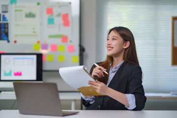 Young Asian business professional smiling, holding a clipboard and pen, taking notes in a modern office environment