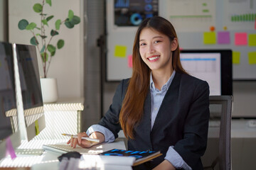 Asian businesswoman confidently smiling, sitting at desk, working on computer in a professional office environment