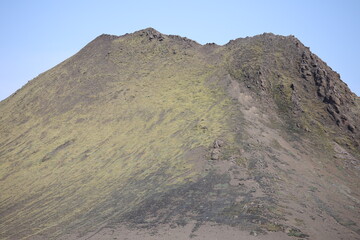 Rugged, moss-covered volcanic mountain slope under a clear blue sky, showcasing raw, untamed...