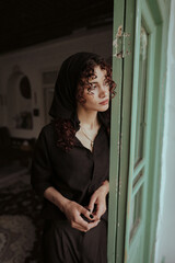 Fototapeta premium Portrait of a Persian woman with curly hair standing by a green wooden door, wearing black clothes and headscarf – emotional fine art indoor photography in soft natural light