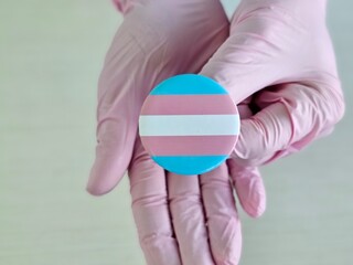 Hands in gloves hold a circular pin featuring the transgender pride flag