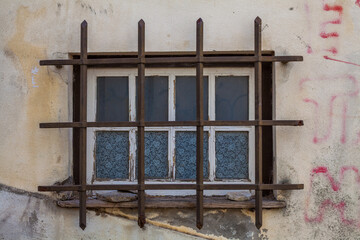 A weathered window with peeling white paint and patterned glass is secured by a rusty metal grille