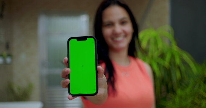 Woman holding smartphone with green screen chroma key close to the camera, smiling in an outdoor home environment, showcasing a blank screen for customizable content