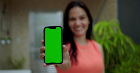 Woman holding smartphone with green screen chroma key close to the camera, smiling in an outdoor home environment, showcasing a blank screen for customizable content