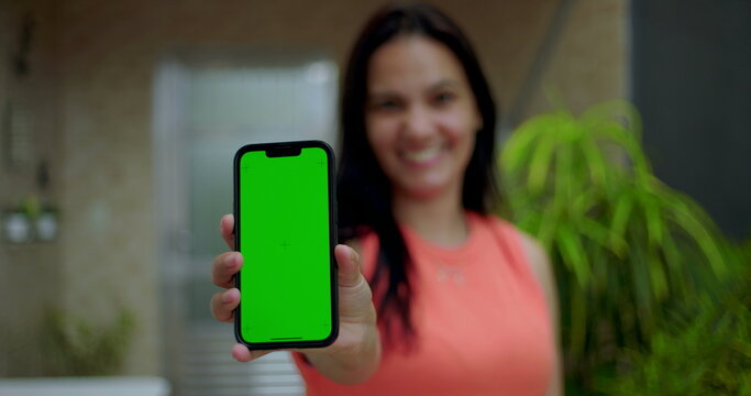 Woman holding smartphone with green screen chroma key close to the camera, smiling in an outdoor home environment, showcasing a blank screen for customizable content - Powered by Adobe