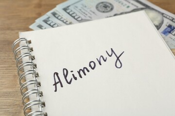 Notebook with word alimony and money on wooden table, closeup