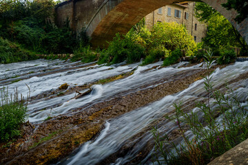 Fast flowing river under old bridge, greenery and historic stone building