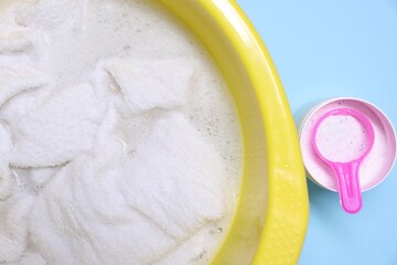 Plastic basin with clothes and washing powder on light blue background, top view