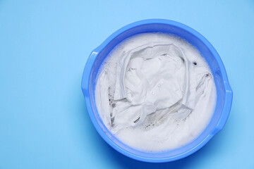 Plastic basin with clothes, water and foam on light blue background, top view