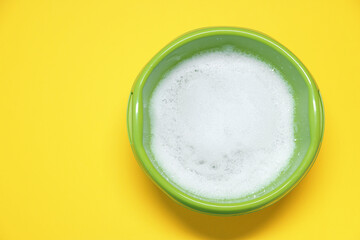 Green plastic basin with water and foam on yellow background, top view. Space for text