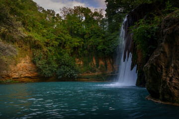 Obraz premium Tall natural waterfall plunging to blue river pool, surrounded by lush forest cliffs
