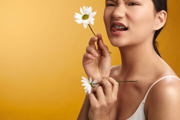 Fototapeta premium asian woman holds a delicate flower near her face, smiling softly against a warm yellow backdrop, conveying a fresh and gentle mood that highlights natural beauty and calm confidence.